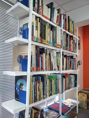 A well-organized bookshelf filled with a diverse collection of books arranged in various colors and sizes. The shelves are white and made of metal, contributing to a clean and modern look. Various blue bookends with a globe design are used to support the books, and the wall beside the shelf is painted in a warm, orange tone.