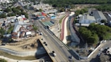 Aerial view of an urban area featuring a road with several vehicles, adjacent to a canal or drainage system with concrete walls. On one side of the road, there is a residential neighborhood with densely packed houses and the other side has commercial and industrial buildings. The area is surrounded by lush green vegetation.