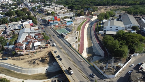 Aerial view of an urban area featuring a road with several vehicles, adjacent to a canal or drainage system with concrete walls. On one side of the road, there is a residential neighborhood with densely packed houses and the other side has commercial and industrial buildings. The area is surrounded by lush green vegetation.