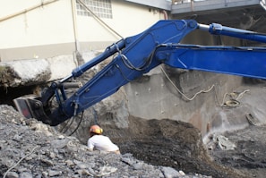 A construction site featuring a large blue excavator digging into the ground near a concrete wall. There is a worker wearing a helmet and noise-cancelling earmuffs near the excavator observing the work. The ground is covered with dark dirt and debris. In the background, there are concrete walls and part of a yellow building with a window and some pipes.