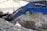A construction site featuring a large blue excavator digging into the ground near a concrete wall. There is a worker wearing a helmet and noise-cancelling earmuffs near the excavator observing the work. The ground is covered with dark dirt and debris. In the background, there are concrete walls and part of a yellow building with a window and some pipes.