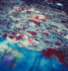 A detailed texture of fabric with an intricate floral pattern. The fabric features various shades of blue and red, with flowers delicately scattered across the surface. Light and shadow create a dynamic, soft-focus effect, adding depth and visual interest.