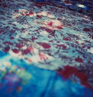 A detailed texture of fabric with an intricate floral pattern. The fabric features various shades of blue and red, with flowers delicately scattered across the surface. Light and shadow create a dynamic, soft-focus effect, adding depth and visual interest.