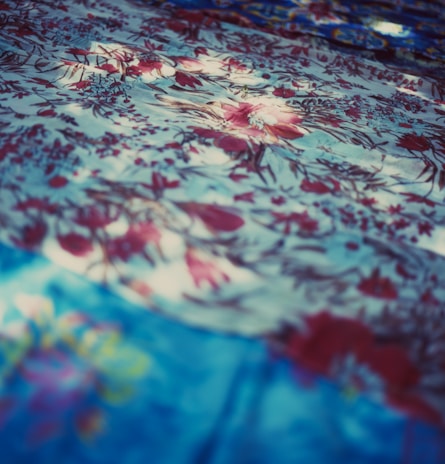 A detailed texture of fabric with an intricate floral pattern. The fabric features various shades of blue and red, with flowers delicately scattered across the surface. Light and shadow create a dynamic, soft-focus effect, adding depth and visual interest.