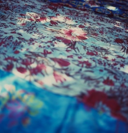 A detailed texture of fabric with an intricate floral pattern. The fabric features various shades of blue and red, with flowers delicately scattered across the surface. Light and shadow create a dynamic, soft-focus effect, adding depth and visual interest.