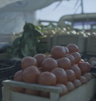 A crate filled with ripe red tomatoes is in the foreground. The setting appears to be a market stall with other vegetables and crates in the blurred background, suggesting a rustic and fresh atmosphere.