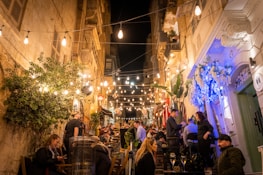 A lively alleyway filled with people sitting at tables, adorned with string lights that create a warm and inviting atmosphere. The architecture is classical, with stone buildings and ornate details. Lush greenery and vibrant flowers accentuate the scene, suggesting a charming European street.