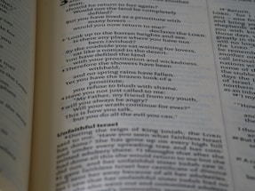 A close-up view of an open book displaying text written in a poetic format across two pages. The left page focuses on verse-style text with religious language, possibly from a scripture. The right page has more formal prose, with headings such as 'Unfaithful Israel'. The pages are cream-colored with dark text and a faint blue tint.