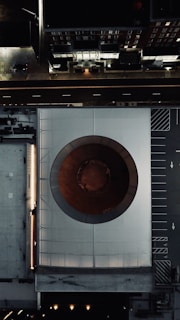 An aerial view of a modern architecture building with a large circular opening in the center, surrounded by urban infrastructure including roads and parking spaces. The image captures a blend of geometric patterns and urban design elements.