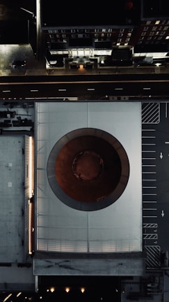 An aerial view of a modern architecture building with a large circular opening in the center, surrounded by urban infrastructure including roads and parking spaces. The image captures a blend of geometric patterns and urban design elements.