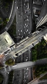 An aerial view of a complex urban highway interchange with multiple lanes of traffic. The image reveals several cars densely packed on the roadways, surrounded by tall buildings and patches of greenery. The road infrastructure includes overpasses and intersecting lanes.