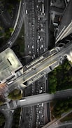An aerial view of a complex urban highway interchange with multiple lanes of traffic. The image reveals several cars densely packed on the roadways, surrounded by tall buildings and patches of greenery. The road infrastructure includes overpasses and intersecting lanes.