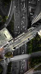 An aerial view of a complex urban highway interchange with multiple lanes of traffic. The image reveals several cars densely packed on the roadways, surrounded by tall buildings and patches of greenery. The road infrastructure includes overpasses and intersecting lanes.