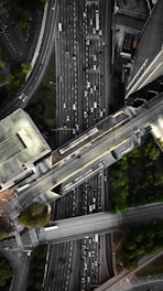 An aerial view of a complex urban highway interchange with multiple lanes of traffic. The image reveals several cars densely packed on the roadways, surrounded by tall buildings and patches of greenery. The road infrastructure includes overpasses and intersecting lanes.