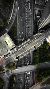 An aerial view of a complex urban highway interchange with multiple lanes of traffic. The image reveals several cars densely packed on the roadways, surrounded by tall buildings and patches of greenery. The road infrastructure includes overpasses and intersecting lanes.