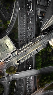 An aerial view of a complex urban highway interchange with multiple lanes of traffic. The image reveals several cars densely packed on the roadways, surrounded by tall buildings and patches of greenery. The road infrastructure includes overpasses and intersecting lanes.