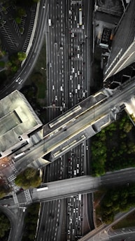 An aerial view of a complex urban highway interchange with multiple lanes of traffic. The image reveals several cars densely packed on the roadways, surrounded by tall buildings and patches of greenery. The road infrastructure includes overpasses and intersecting lanes.