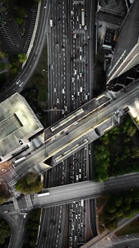 An aerial view of a complex urban highway interchange with multiple lanes of traffic. The image reveals several cars densely packed on the roadways, surrounded by tall buildings and patches of greenery. The road infrastructure includes overpasses and intersecting lanes.