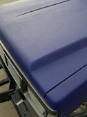 A close-up view of a matte textured blue car hood with part of the front grille and headlight visible. The surface appears even and non-reflective, indicating a durable finish.
