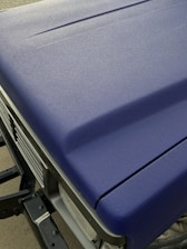A close-up view of a matte textured blue car hood with part of the front grille and headlight visible. The surface appears even and non-reflective, indicating a durable finish.