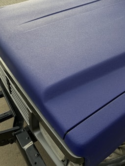 A close-up view of a matte textured blue car hood with part of the front grille and headlight visible. The surface appears even and non-reflective, indicating a durable finish.