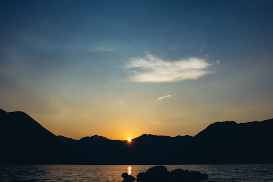 A serene sunset over a tranquil body of water with silhouetted mountains in the background. The sky transitions from deep blue to a warm orange hue near the horizon.