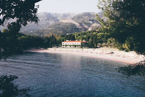 A tranquil beach scene with calm water along a deserted sandy shore. A large building with a red roof sits nestled among lush greenery and tall trees. Mountains rise in the background, adding depth to the landscape. The overall setting is peaceful and undisturbed, with shadows from overhanging branches framing the view.