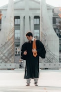 A person wearing a graduation gown and a gold stole stands in an open space in front of a monumental structure with tall columns and artistic designs. The individual appears to be adjusting their attire while looking down.