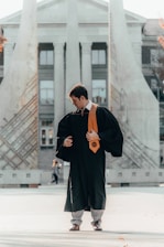 A person wearing a graduation gown and a gold stole stands in an open space in front of a monumental structure with tall columns and artistic designs. The individual appears to be adjusting their attire while looking down.