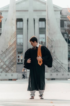 A person wearing a graduation gown and a gold stole stands in an open space in front of a monumental structure with tall columns and artistic designs. The individual appears to be adjusting their attire while looking down.