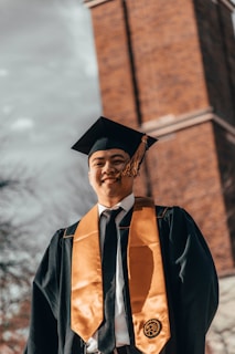 A person is dressed in a graduation cap and gown, standing outdoors with a tall brick tower in the background. The gown is dark with an orange stole and the person is smiling.
