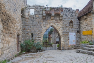 A historical stone wall with an arched gateway featuring a metal grate. The structure includes weathered bricks and small plants growing in crevices. An informational poster is displayed on the right side of the arch, and there are some stairs leading up to a higher level with a small garden area.