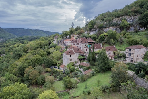 A quaint village nestled among lush, rolling hills with a collection of rustic, stone houses with red-tiled roofs. The buildings are surrounded by abundant greenery, including trees and carefully tended gardens. A rocky hillside lies behind the village, adding to the secluded and serene atmosphere.