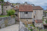 A picturesque medieval village scene with stone houses featuring timber framing and steep, gabled roofs. The narrow cobblestone path leads downward past an ancient stone wall, surrounded by lush greenery. The architecture is characteristic of a historical European rural setting.