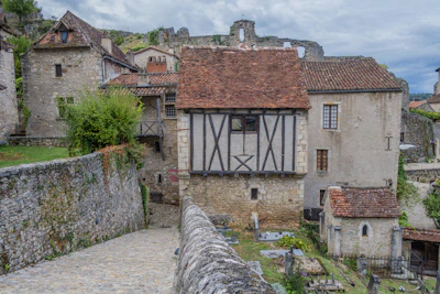 A picturesque medieval village scene with stone houses featuring timber framing and steep, gabled roofs. The narrow cobblestone path leads downward past an ancient stone wall, surrounded by lush greenery. The architecture is characteristic of a historical European rural setting.