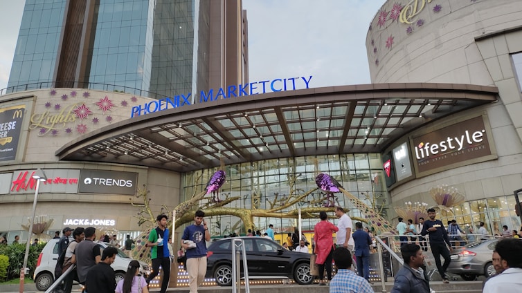 A bustling shopping mall entrance with people walking around. The architecture features a large glass facade and several brand logos, including Jack & Jones, Lifestyle, and others. Decorative elements like artificial trees and flowers add a festive touch. Cars are parked nearby, contributing to a dynamic urban scene.
