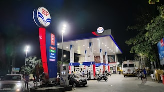 A well-lit fuel station at night, featuring multiple fuel pumps and a prominent HP logo. Vehicles, including cars and motorcycles, are present at the pumps. The area is illuminated by tall lights, and there are trees in the vicinity.