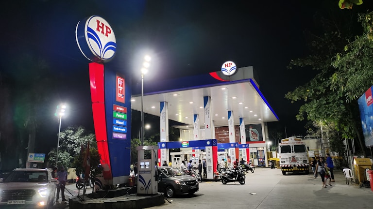 A well-lit fuel station at night, featuring multiple fuel pumps and a prominent HP logo. Vehicles, including cars and motorcycles, are present at the pumps. The area is illuminated by tall lights, and there are trees in the vicinity.