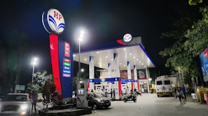 A well-lit fuel station at night, featuring multiple fuel pumps and a prominent HP logo. Vehicles, including cars and motorcycles, are present at the pumps. The area is illuminated by tall lights, and there are trees in the vicinity.