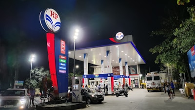 A well-lit fuel station at night, featuring multiple fuel pumps and a prominent HP logo. Vehicles, including cars and motorcycles, are present at the pumps. The area is illuminated by tall lights, and there are trees in the vicinity.
