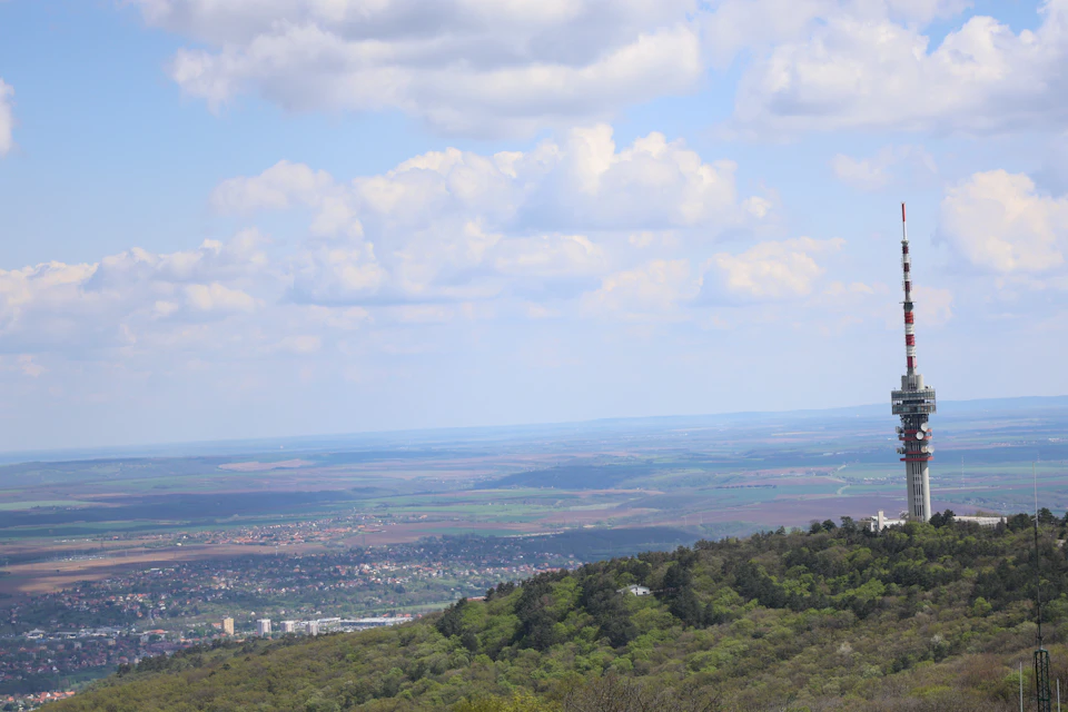The image depicts a serene landscape with a tall, white radio tower situated on a hilltop. The tower is surrounded by lush greenery and features two antennae extending from its top. In the distance, there are several buildings scattered across the horizon line, suggesting that this location may be in an urban area or near a city center. The sky above is filled with clouds, casting a soft light over the scene and creating a sense of tranquility.