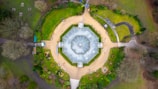 An aerial view of a large, symmetrical glass greenhouse set within a landscaped garden. The greenhouse is octagonal with a transparent roof, surrounded by pathways and green lawns. Various plant beds and walkways are scattered around the structure, bordered by well-maintained greenery and trees.