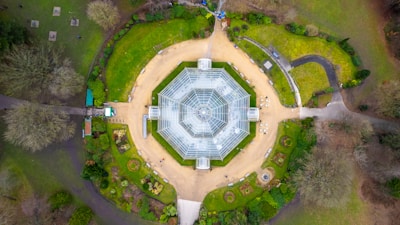 An aerial view of a large, symmetrical glass greenhouse set within a landscaped garden. The greenhouse is octagonal with a transparent roof, surrounded by pathways and green lawns. Various plant beds and walkways are scattered around the structure, bordered by well-maintained greenery and trees.