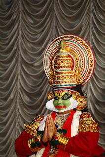 An individual is dressed in a traditional costume, featuring intricate face paint and an elaborate headdress adorned with gold and red details. The hands are clasped together in a gesture resembling a respectful greeting or prayer. The background consists of draped fabric with a silver sheen.