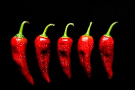 Five vibrant red chili peppers with green stems are arranged in a row against a solid black background, with visible droplets of water on their surfaces, giving them a fresh and glossy appearance.