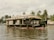 A traditional houseboat with a thatched roof floats on calm waters. The boat has circular windows and a spacious open area. In the background, lush palm trees line the riverbank, and a few birds soar in the sky.