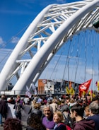 A large group of people is gathered on a bridge with a distinct white, modern arch design. The crowd is holding various colorful flags, some with visible text. A clear, blue sky is in the background, and there are buildings visible in the distance. A traffic sign indicating a speed limit of 50 is present.