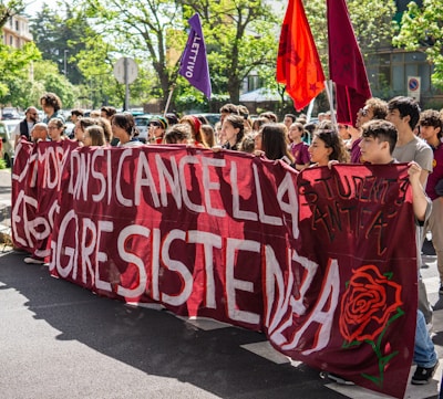 A group of people, mostly young adults, are gathered together on a street holding large banners with slogans. The banners are predominantly red with white and black text. There are also some flags in purple and red with writing. The people appear to be participating in a protest or demonstration. The background shows trees and buildings, suggesting an urban setting.