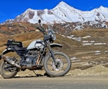 A motorcycle is parked on a road in front of a mountainous landscape. The mountain background has patches of snow at the peaks and a few scattered buildings are visible in the mid-ground. The sky is clear and blue, adding a vibrant backdrop to the scene. The motorcycle has dirt and signs of usage, indicating off-road travel.