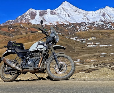 A motorcycle is parked on a road in front of a mountainous landscape. The mountain background has patches of snow at the peaks and a few scattered buildings are visible in the mid-ground. The sky is clear and blue, adding a vibrant backdrop to the scene. The motorcycle has dirt and signs of usage, indicating off-road travel.